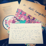 A photograph of three print copies of issue one of Doll Hospital Journal with a handwritten postcard placed on the top