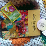 A photograph of Doll Hospital Issue One, a lollipop, two doll hospital stickers and a Pokemon card of Koffing on a colourful crochet blanket